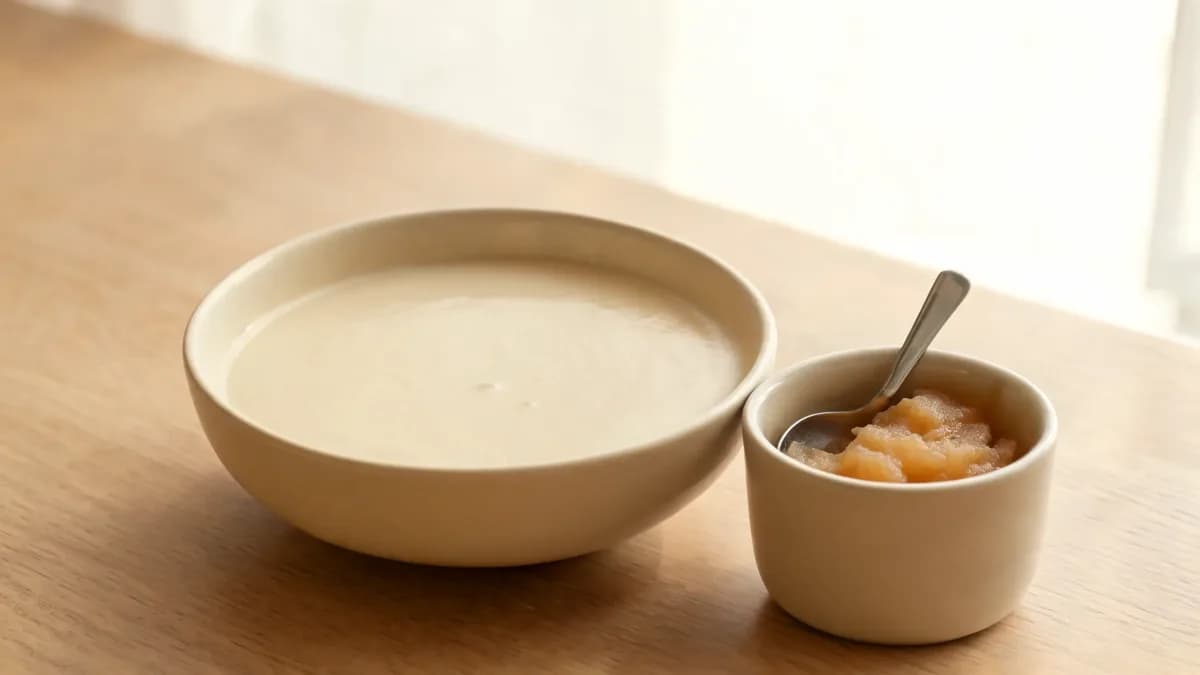 A bowl of lukewarm creamy soup and a cup of applesauce with a spoon on a light table